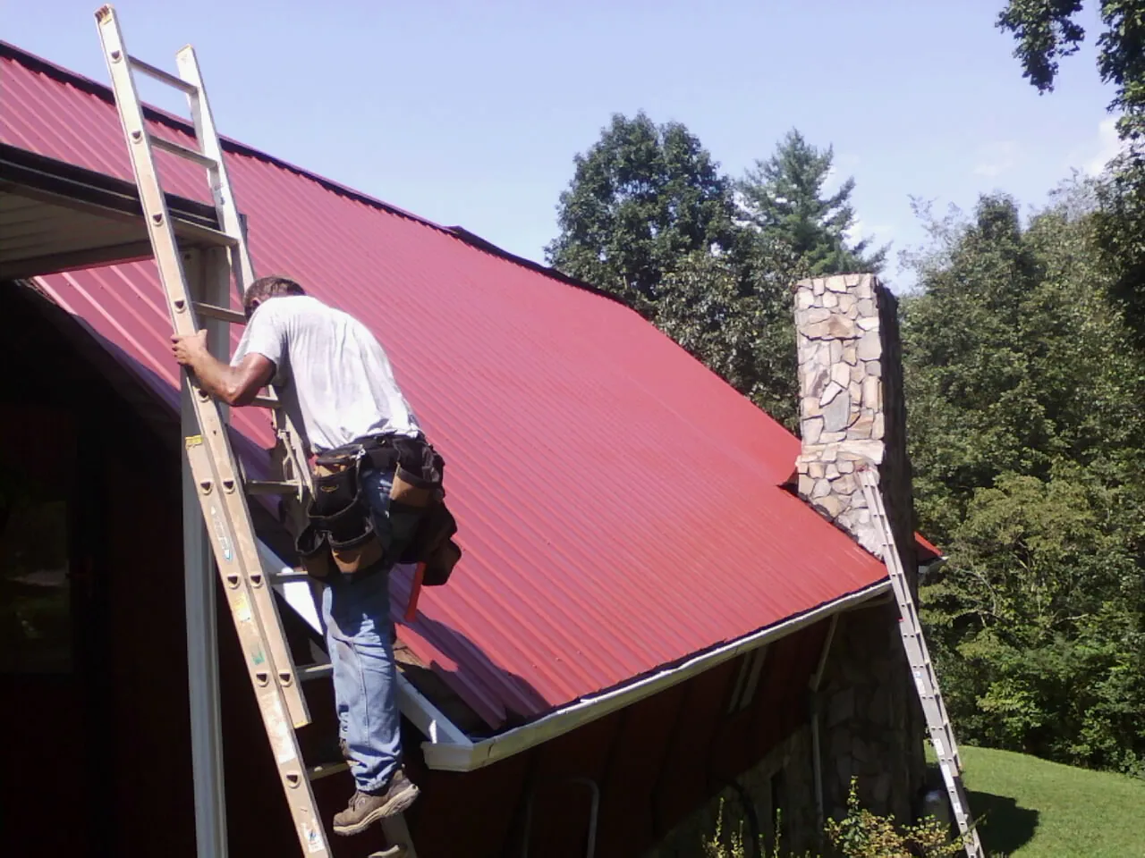 Roofer on a ladder beside a red metal roof and stone chimney — Soffit & Fascia Repair by Roofing Brothers in Fargo